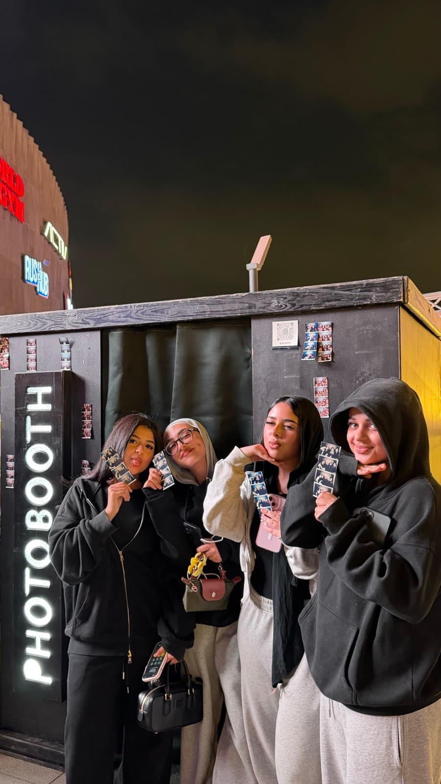 Four guests holding their printed photo strips beside the black Tru Booth photo booth at night